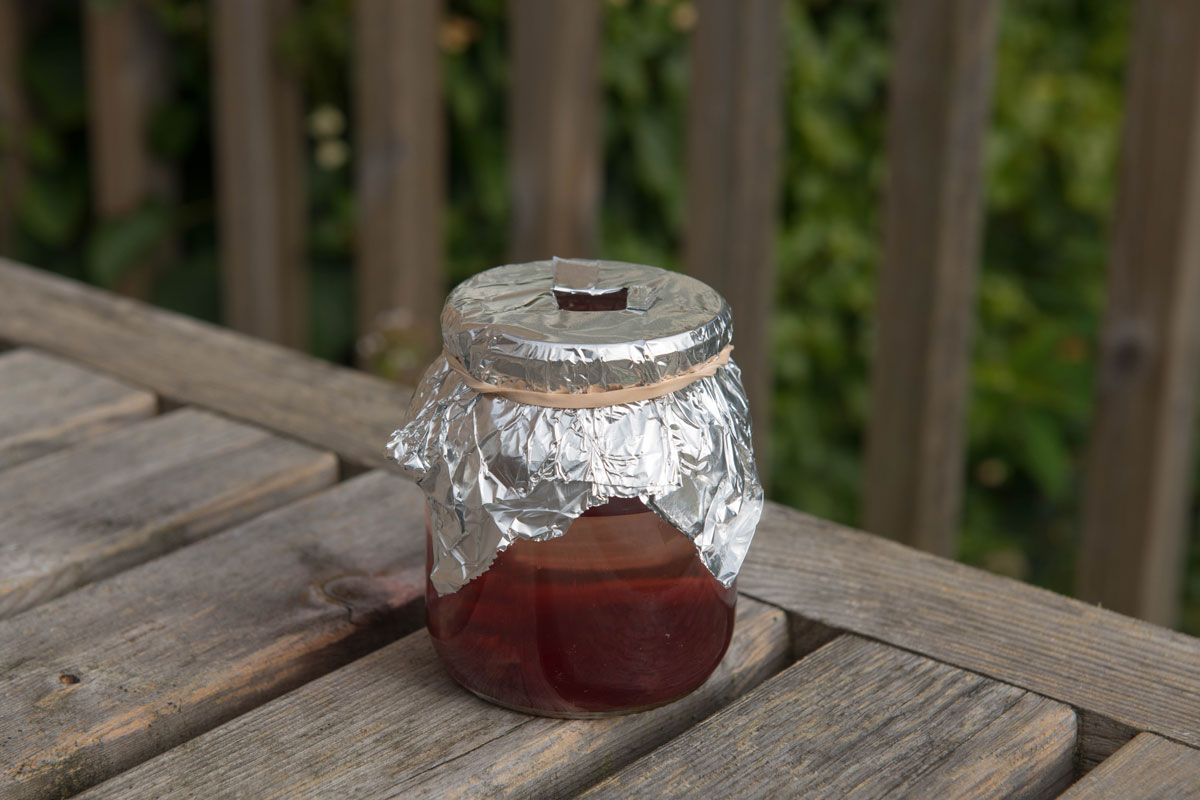 Home Made Wasp Trap from a Glass Jar Filled with Jam and Water Secured with Silver Foil and a Rubber Band on an Outdoor Table Top